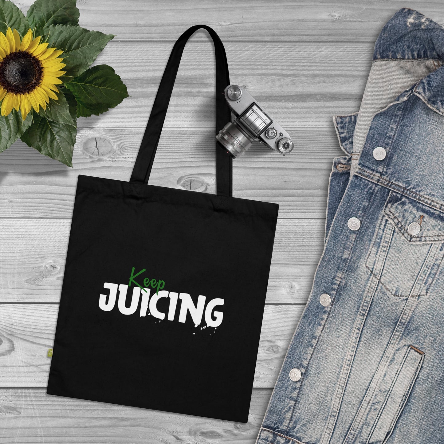 Black tote bag with 'Keep Juicing' text on a wooden surface with a sunflower and denim jacket.