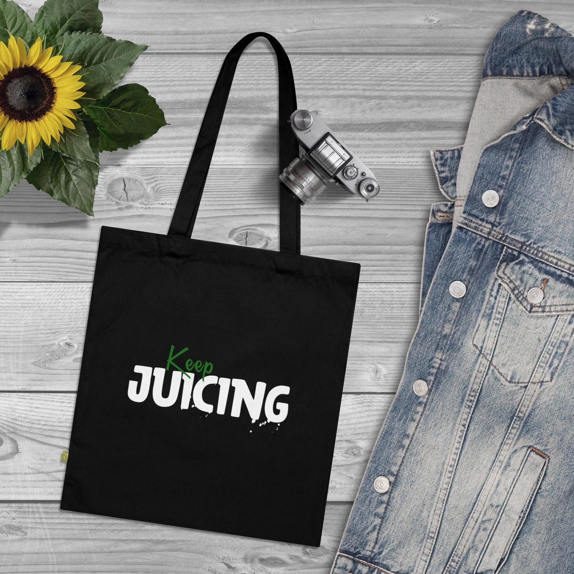 Black tote bag with 'Keep Juicing' text on a wooden surface with a sunflower and denim jacket.